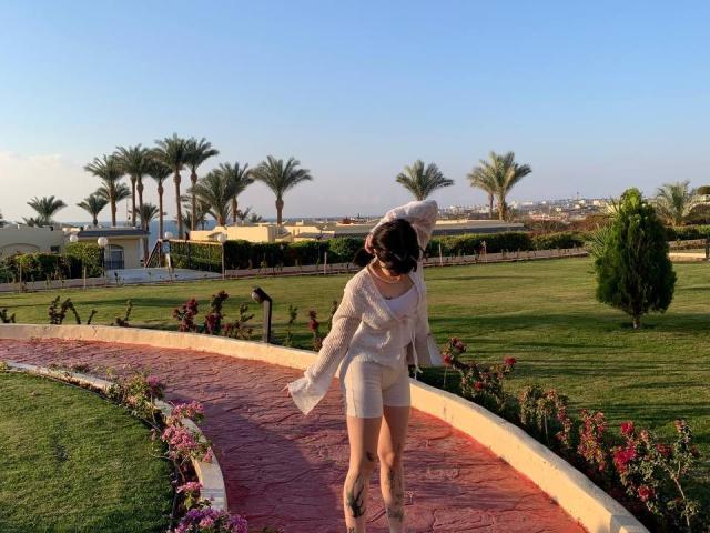 LovelDiva in a white outfit and hat, blowing her hair, standing on a red brick path with green grass and palm trees, wearing beige boots.