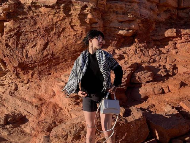 LovelDiva with black hair, glasses, black sweater, checkered jacket, black shorts, holding white bag, standing on rocky red-orange terrain. Her tits are small, she's cute.