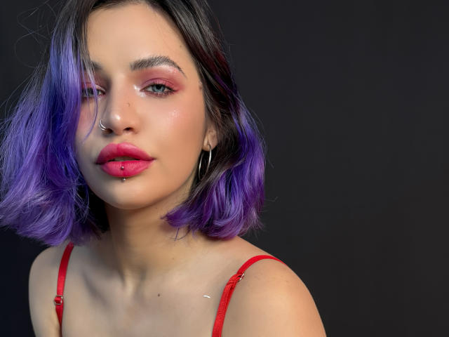 BriannaAngel69 has purple hair, pink eyeshadow, red lipstick with a lip ring, nose ring, and hoop earrings, wearing a red bra, against a dark background.