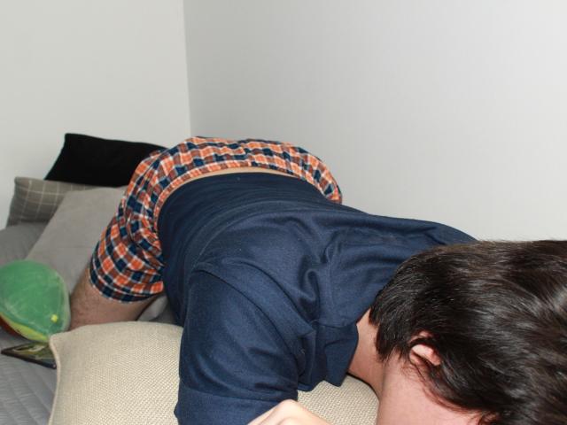 ErossGuy is on a bed, knees and elbows down, butt up, wearing orange and blue checkered shorts and a dark navy shirt. His head is down, facing right.