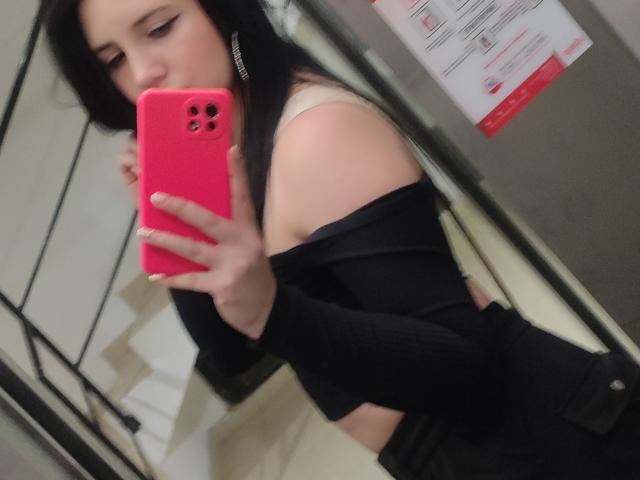 Kkatalina with black hair, pink phone, off-shoulder black top, white bra, holding phone in mirror selfie, showing her bare shoulder and upper chest in a bathroom.