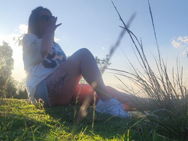 Kkatalina sits on grass, legs crossed, wearing a white tee and denim shorts, sipping from a bottle, sun flares, grass in foreground, tattoos on right thigh, sunglasses on.