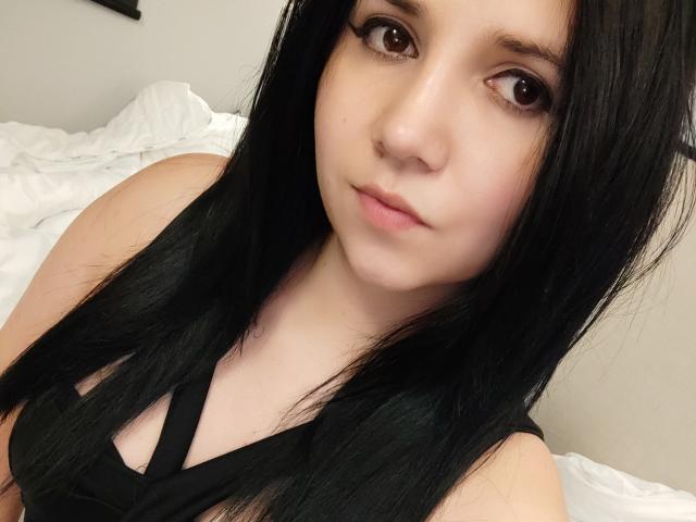 Kkatalina with long black hair, brown eyes, and a neutral expression, wearing a black strappy top, showing her bare shoulders and upper chest, in a selfie.