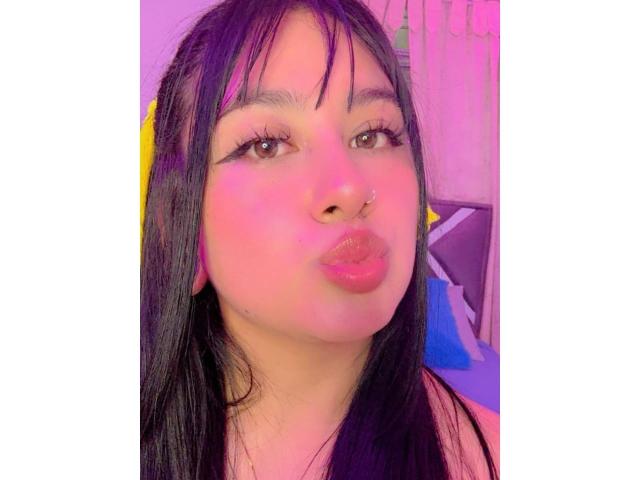AdharaBritney with puffy pink lips, black hair, yellow ear cover, and nose ring, making a pouty face, surrounded by pink and purple lighting.