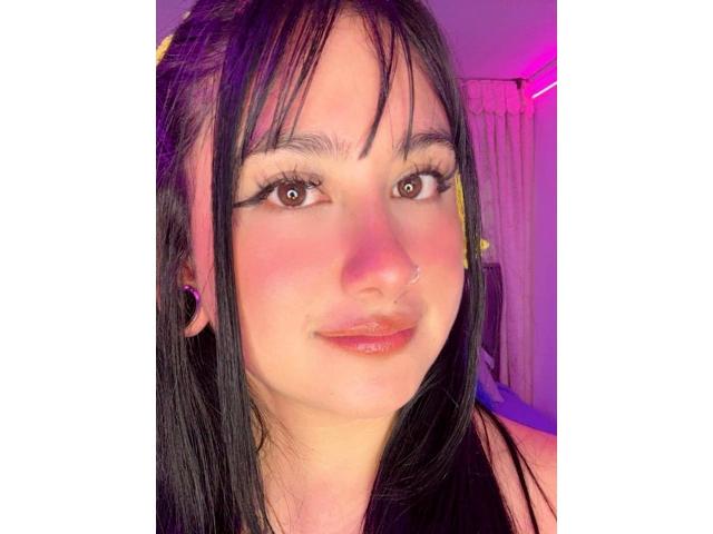 AdharaBritney has black hair, brown eyes, a nose ring, and pinkish cheeks with glossy lips. Her makeup includes bold eyeliner, and she's wearing pink earrings.