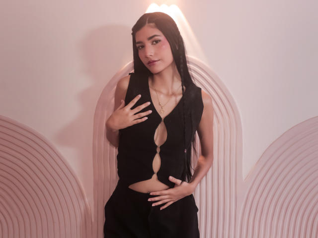 Roxanna Herias, con lunghi capelli neri, indossa un abito nero con aperture, mani sul petto e sull'addome, luce rosa tenue, parete ad arco semi-trasparente sullo sfondo, posa sexy e sicura di sé..