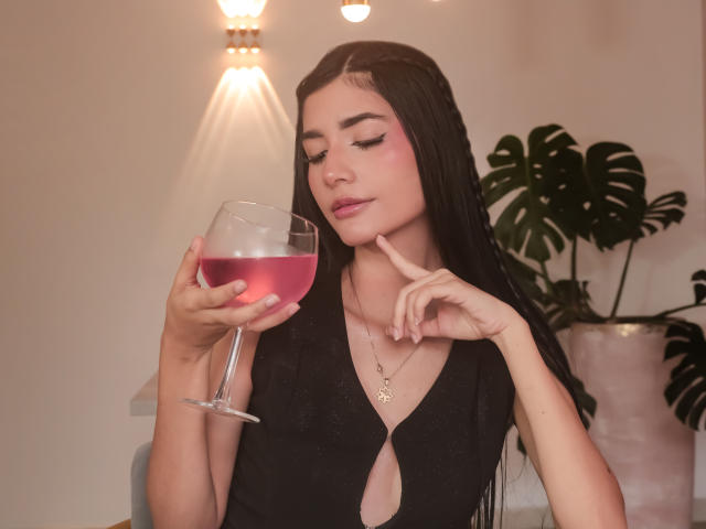 RoxannaHerias, com cabelos longos e pretos, blush rosa, vestido preto, segurando uma taça de vinho rosa, com o dedo no queixo, usando um colar, com um sorriso discreto, lábios carnudos, iluminação suave, em um ambiente interno..