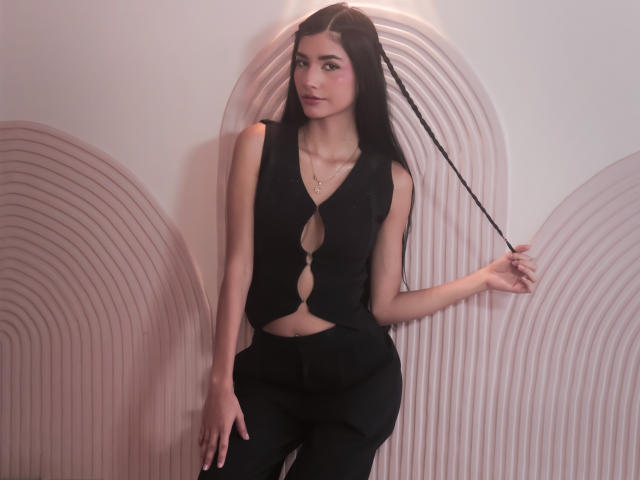 RoxannaHerias with long black hair, wearing a black cut-out vest and pants, standing, holding a braid, slim, pale skin, subtle makeup, pendant necklace, sexy, confident pose.