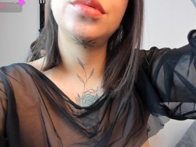 KayleeM's face, showing her lips slightly parted, with a tattoo of a rose and leaves on her chest, wearing a sheer black top, brown hair. Boobs not visible.