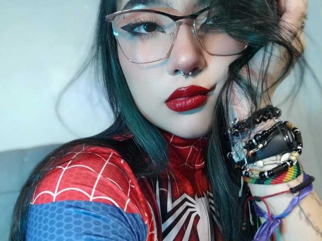 LyRose with dark green hair, red lipstick, nose ring, glasses, and Spider-Man costume, showing off colorful wrist bracelets, looking at camera with a slight smile.