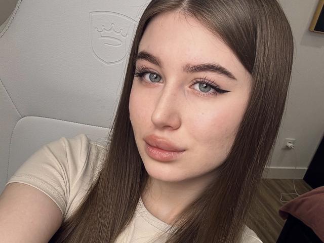 LillyColins, con cabello castaño largo, ojos azules y delineador de ojos alado, vestida con una camisa beige, sentada en una silla blanca, mirando directamente a la cámara con una ligera sonrisa..