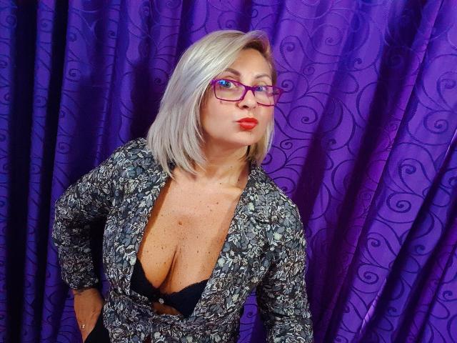 ChaudeEvely with purple glasses, red lipstick, and gray patterned top showing black bra, revealing her boobs. She has silver hair and a confident expression.