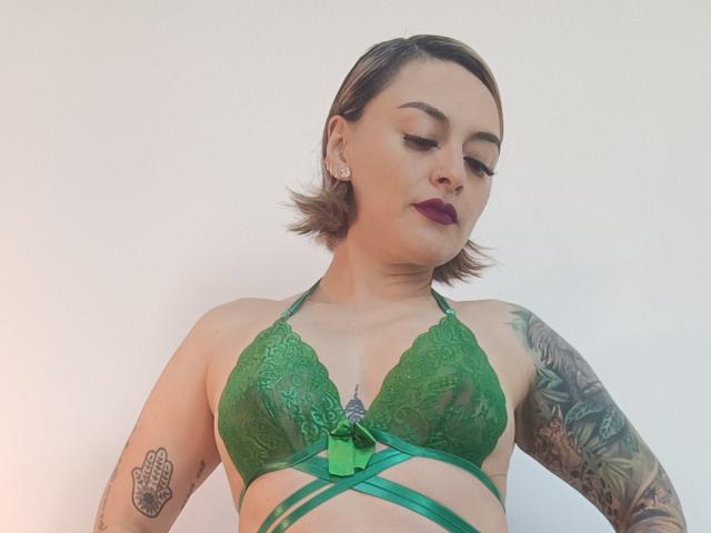 KatParadise, con cabello corto y castaño, un sujetador de encaje verde con lazo, lápiz labial morado, tatuajes en el brazo izquierdo y el pecho derecho, mirando hacia abajo, con un fondo de pared blanca..