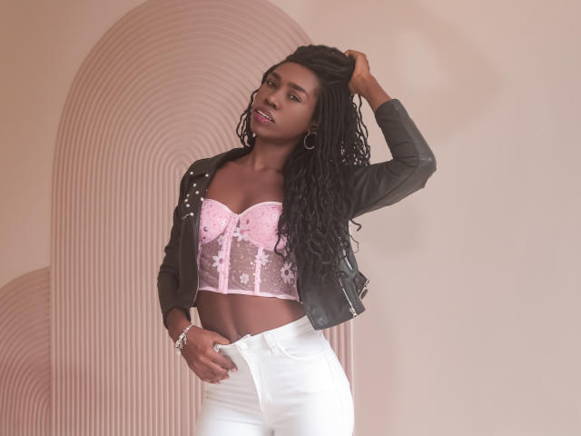 SofiaKem with dark skin, long black dreadlocks, wearing a pink lace-up bustier, black jacket, and white pants, hand in hair, looking at camera, pink lipstick, hoop earrings.