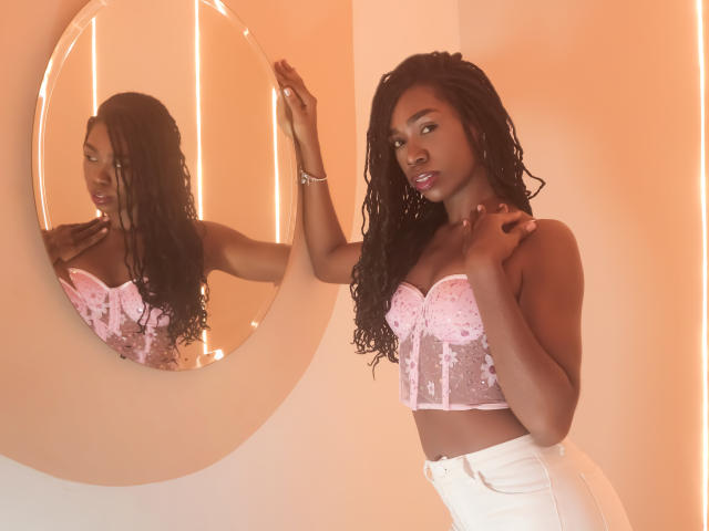 SofiaKem with dark skin, long curly black hair, pink lace-up top, showing her tits, white high-waisted pants, standing, touching round mirror, soft orange lighting, reflective mirror.