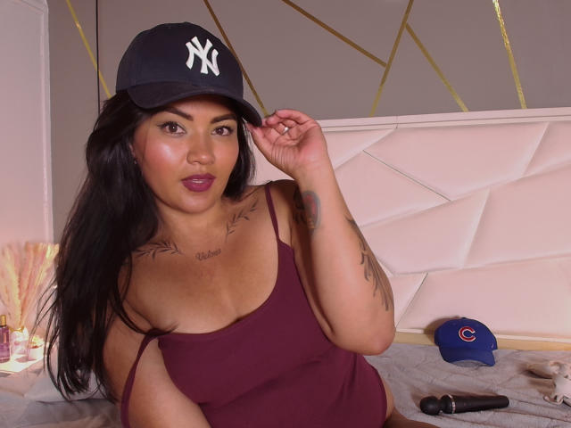 IvySoul wearing a black NY Yankees cap, purple tank top, showing tattoos on chest and arm, sitting on bed with a blue cap and black wand on right.
