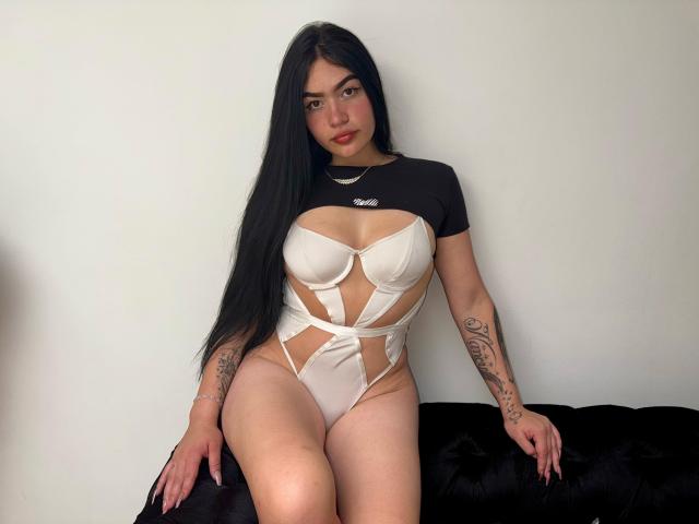AdeleMontana with long black hair, wearing a white cut-out bodysuit, exposing her big tits and showing tattoos on her arms, sitting on a black furry surface.