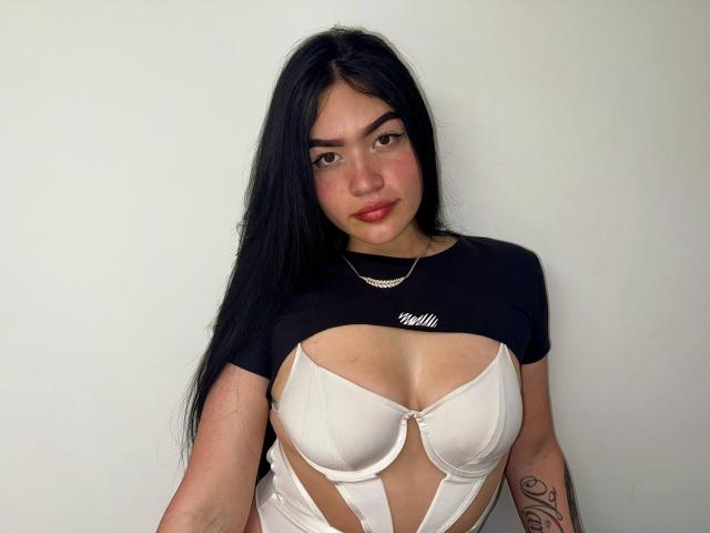 AdeleMontana with long black hair, wearing a black top and white cut-out bra exposing her big tits, standing against a white wall, looking at the camera.