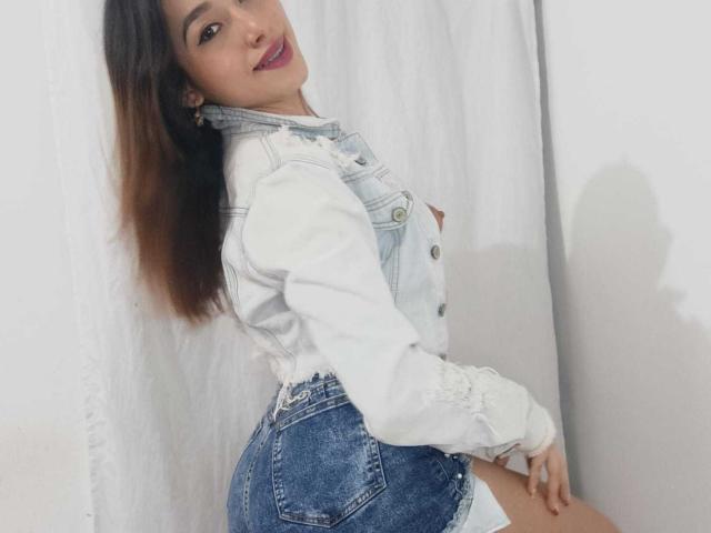 Alexa Laker, con lunghi capelli castani, indossa una giacca di jeans bianca e pantaloncini blu a vita alta, mostrando i suoi fianchi formosi, sorride con un rossetto viola, ed è in piedi davanti a una tenda bianca..
