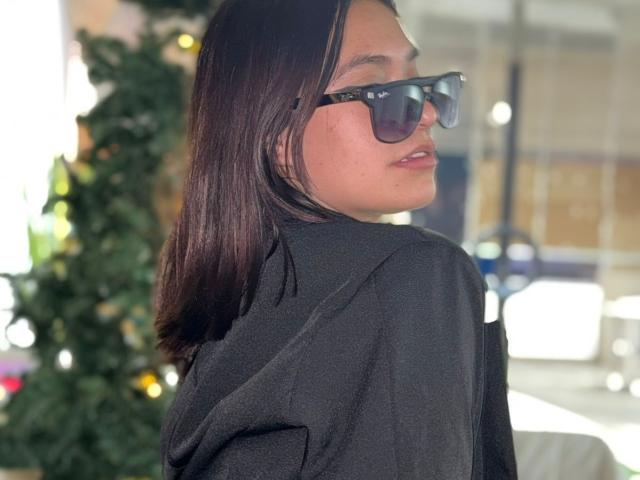 CassieJoness, com cabelo castanho escuro, usando óculos de sol pretos e um moletom preto, olhando por cima do ombro, com os lábios ligeiramente entreabertos, mostrando um toque de batom vermelho..