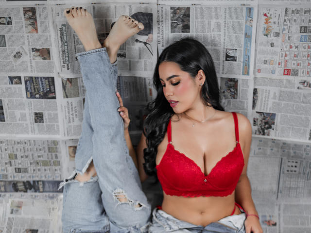 PaolaYKatty with red lace bra, large tits, and ripped jeans, legs up, black nails, long black hair, and pink lipstick, sitting on newspaper.