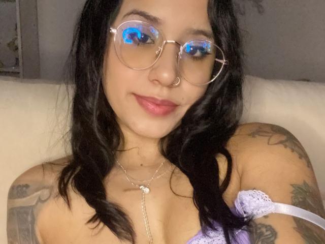 AshleyMayer with blue-tinted glasses, wavy black hair, pink lips, tattoos on arms, purple lace top, showing her tits, smiling, wearing a nose ring, on a white couch.