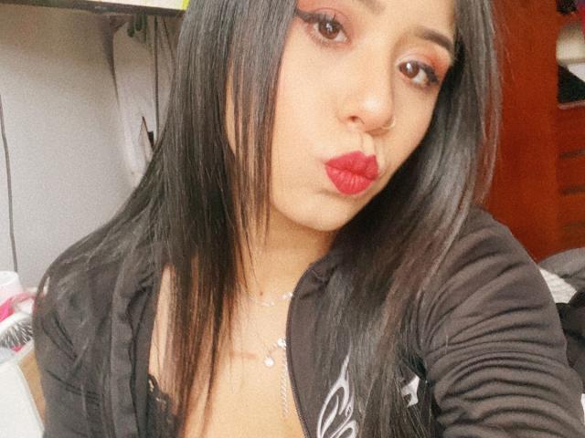 AshleyMayer has long black hair, red lipstick, and is making a pouty face. She's wearing a black shirt with a low neckline, showing some cleavage.