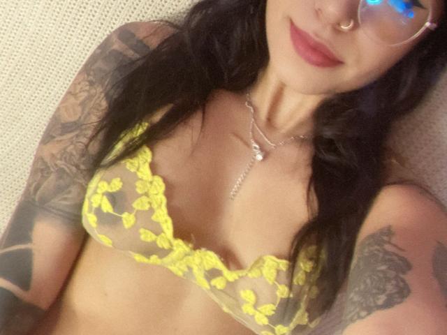 AshleyMayer with yellow lace bra, showing her tits, tattoos on arms, nose ring, glasses, and red lipstick, lying down, smiling.