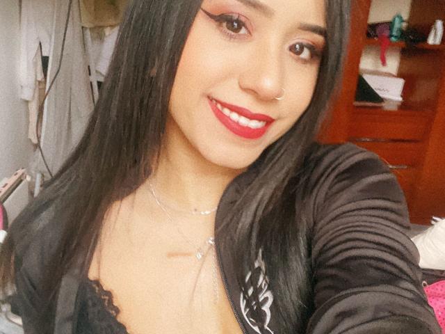 AshleyMayer with long black hair, red lipstick, and black eyeliner, smiling at the camera, wearing a black lace top, and a silver necklace, showing her small tits.