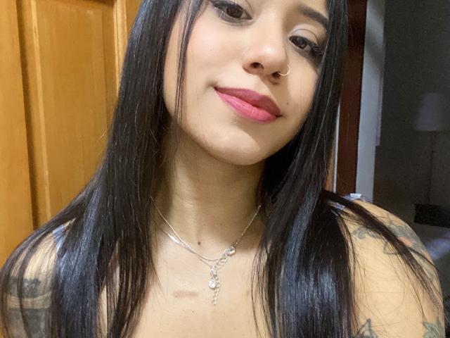 AshleyMayer with long black hair, pink lipstick, nose ring, and tattoos on her arms, wearing a silver necklace, smiling slightly. Her chest is partially visible.