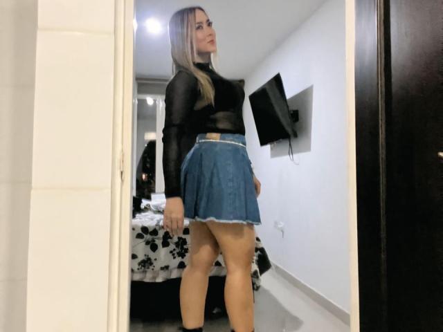 AlinaSmiith stands in a room, wearing a black sheer top and blue denim skirt, showing her ass and a small tan waistband. She's looking back with a slight smile.