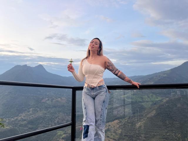 AlinaSmiith stands on a balcony, holding a drink, with one arm resting on the railing. She's wearing a beige off-shoulder top and blue jeans, showing her tits.