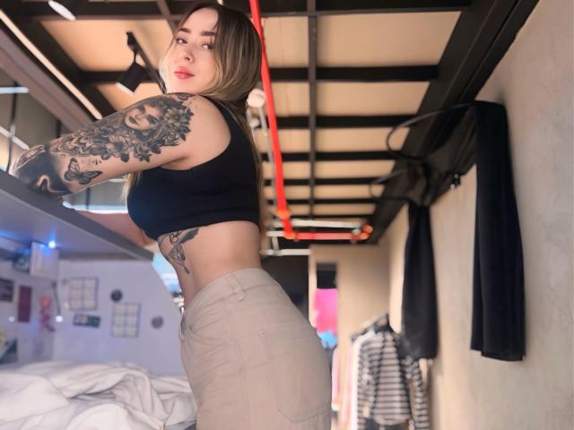 AlinaSmiith with tattoos, black sports bra, and beige pants, showing her ass and tattoos, standing in a room with a black ceiling and red lights.