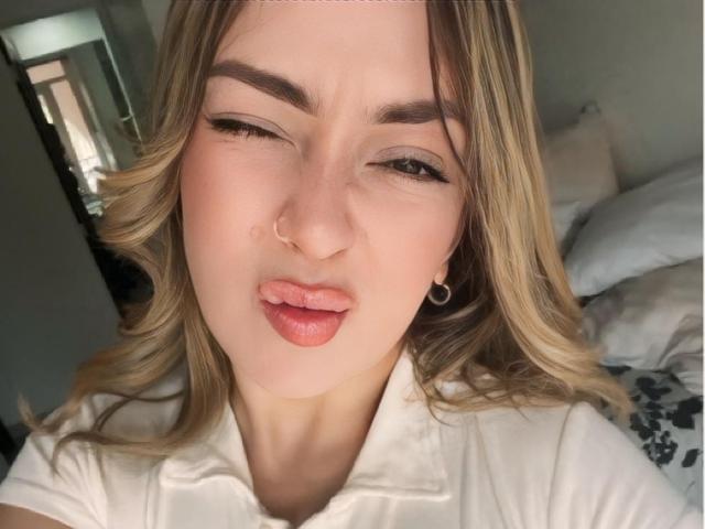 AlinaSmiith's pouty lips are slightly parted, her nose is pierced, and she's winking with one eye, wearing a white shirt, with blonde hair and hoop earrings.