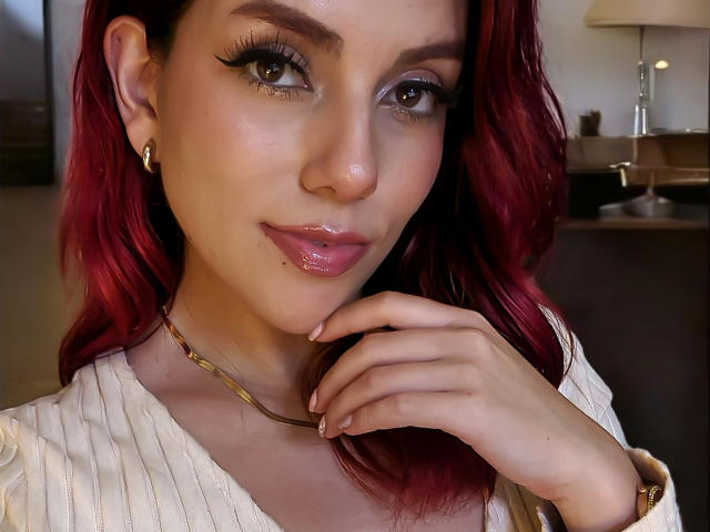 ZoeeMar has shiny red hair, long eyelashes, glossy pink lips, and a gold necklace. She's touching her chin with a hand, wearing a white ribbed shirt.