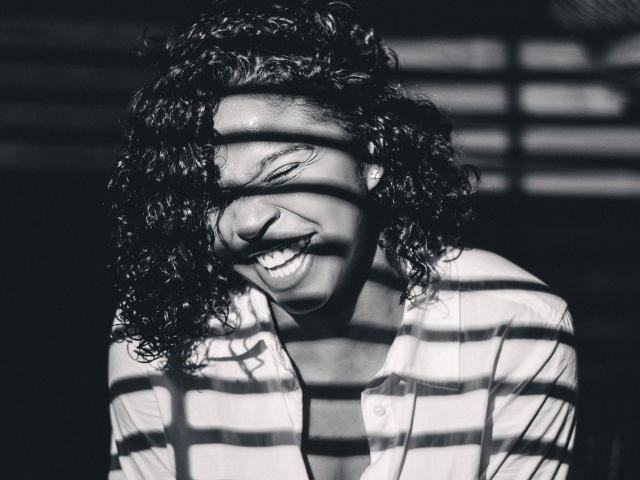 Black-and-white photo of LisaMendez with curly hair, smiling, wearing a striped shirt, shadows from window blinds on her face, showing her teeth and a joyful expression.