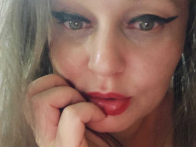 KissAndTits has pale skin, green eyes, and red lipstick. She's biting her finger with black eyeliner, blonde hair, and a slightly parted, sexy mouth. Her hand is near her face.