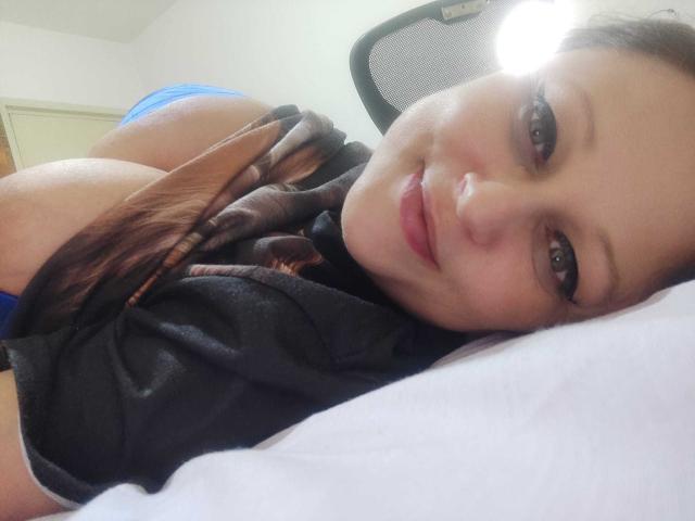 KissAndTits lying on a bed, head resting on white pillow, smiling at camera, brown eyes, black shirt, brown scarf, blue underwear, exposed large boobs, brown hair, soft light.