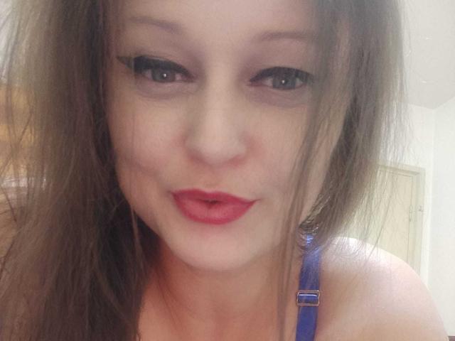 KissAndTits has long brown hair, blue eyes, and red lipstick, wearing a blue strap, with a slightly open mouth and a soft, confident smile.