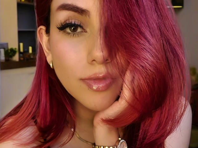 ZoeeMar with shiny pink hair, thick black eyelashes, glossy pink lips, and a hand under her chin, wearing gold earrings and a necklace, has a cute, sexy face.