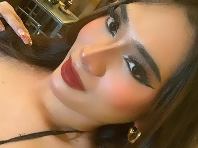 SophiaStarTS has glossy brown eyes, thick black eyebrows, red lipstick, and a slightly flushed face with long dark hair, wearing gold earrings and a black strap.