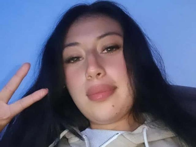 SunnyEve with long black hair, light brown eyes, and pink lips, making a peace sign with her right hand, wearing a beige hoodie, and showing a slight smile.