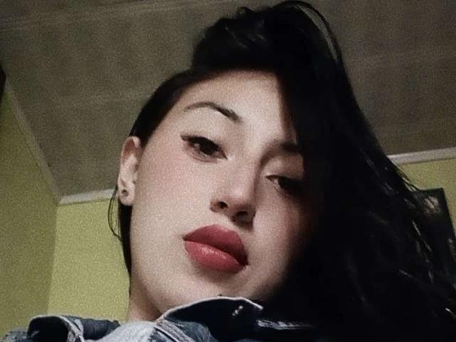 SunnyEve's face, showing dark eyebrows, long black hair, and red lipstick, with a slight downward gaze, wearing a denim jacket, and small hoop earrings, in a low-angle shot.