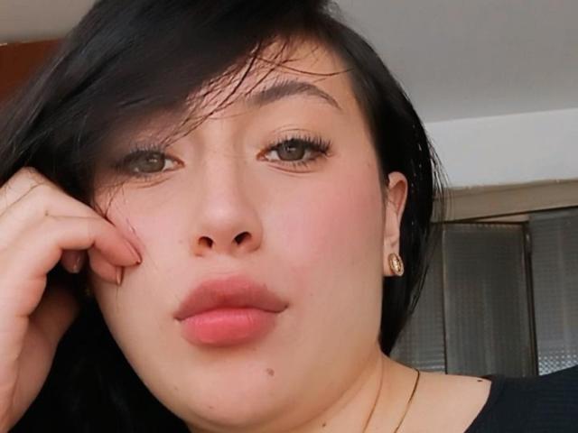 SunnyEve's face with black hair, pink lips, small mole under her nose, green eyes, and gold earrings, touching her cheek. She's wearing a black top and a necklace.