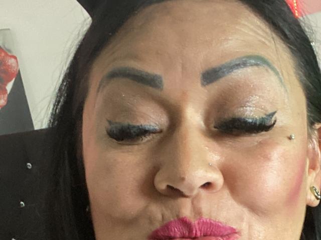 KittyHotLady69 with thick black eyebrows, blue eyeshadow, long eyelashes, nose piercing, and pink lipstick, puckering her lips in a selfie, showing her slightly wrinkled forehead.