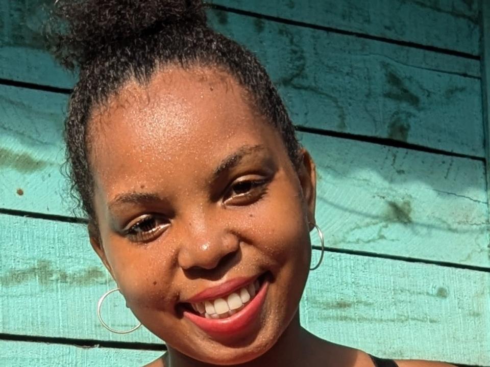 FantasticCute with dark brown skin, curly hair in a top knot, smiling with red lipstick, wearing hoop earrings, against a turquoise wooden background.