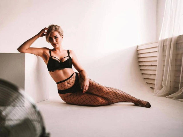 LisaDiamond-hot is laying down, wearing black lingerie and fishnet stockings. She has blonde hair, a slim build, and is looking at the camera with a sultry expression.