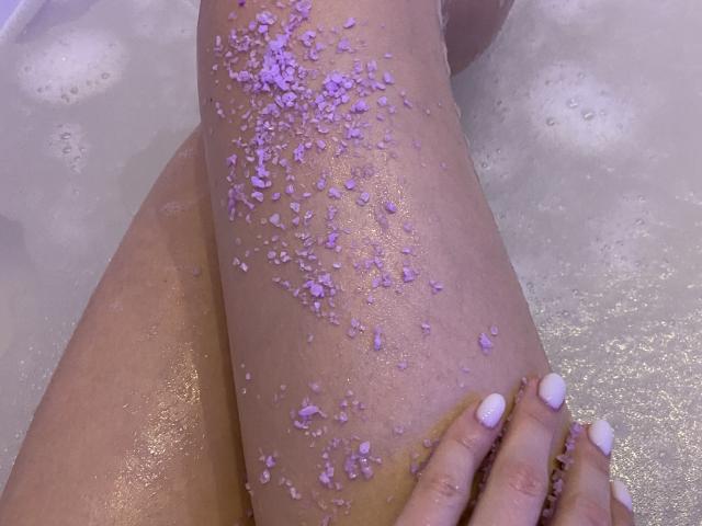PrimeS' legs in a bubble bath, covered in purple salt crystals. PrimeS' hand with white nails rests on the right leg. The water is slightly visible.
