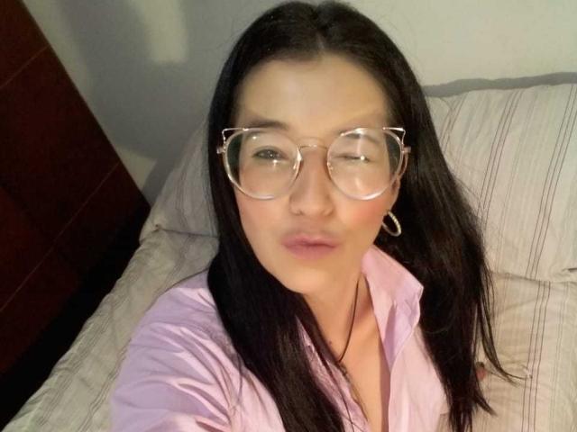 AntonnellaS met een bril, knipend, roze shirt, donker haar, oorringen, liggend op bed, volle roze lippen, zacht licht, licht open roze shirt, bovenlichaam, casual selfie..
