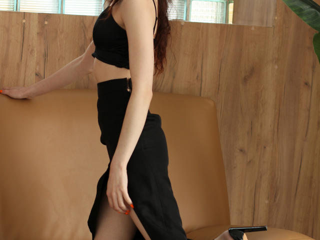 AdrianeWalter with pale skin, red hair, black crop top, and black skirt with a side slit, standing by a brown leather couch, showing her tits and ass.